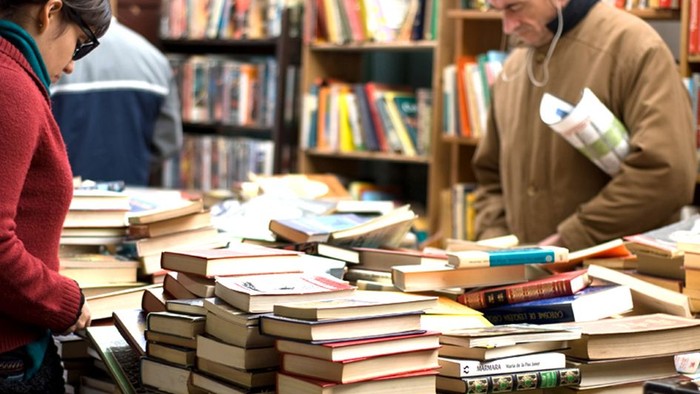 Zwei Personen stöbern in einem Buchladen mit vielen Büchern auf Tischen und Regalen.