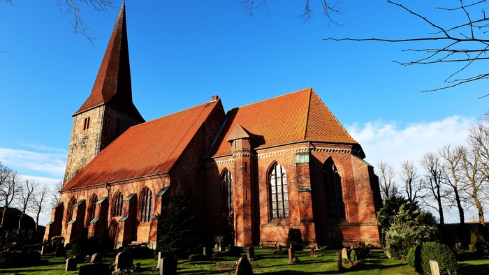Ein rotes Backsteingebäude mit zwei Türmen und einem Friedhof davor unter blauem Himmel