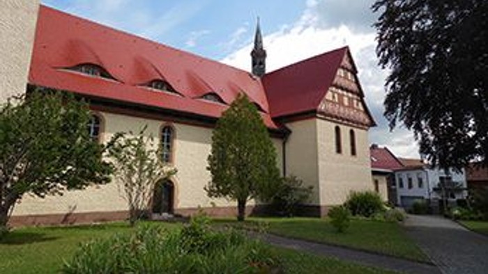 Das Bild zeigt eine Kirche mit rotem Dach und einem Turm, umgeben von Bäumen und einem gepflegten Garten.