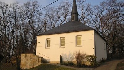 Kleine weiße Kirche mit spitzem Dach und Treppe, umgeben von Bäumen und Gras.