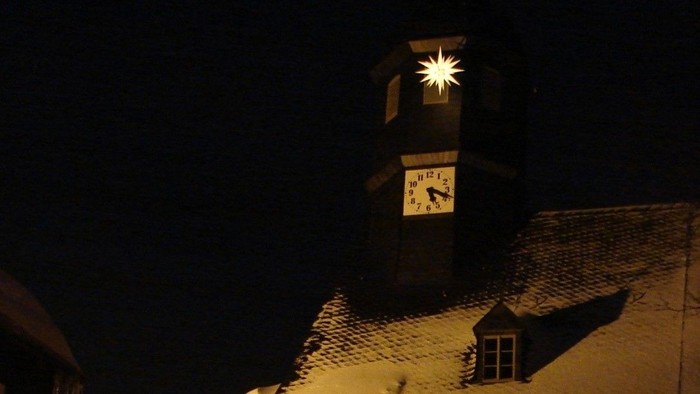 Schneebedeckte Häuser bei Nacht mit beleuchteter Kirchturmspitze