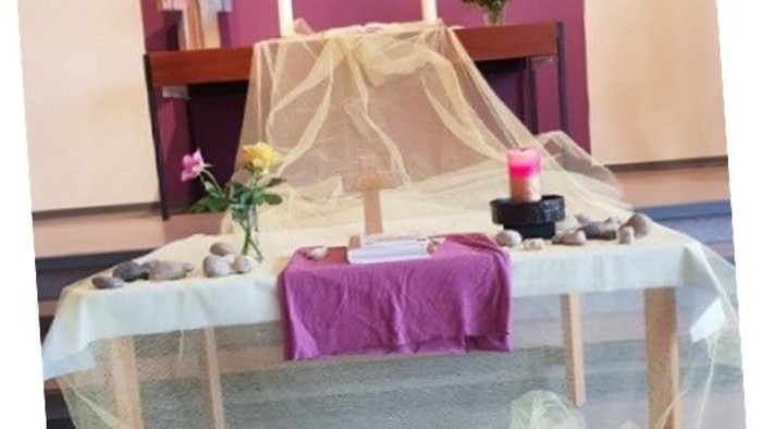 Ein Tisch mit weißer Tischdecke, Blumen und Kerzen, bedeckt mit durchsichtigem Stoff, vor einem lila Tuch und Regalen