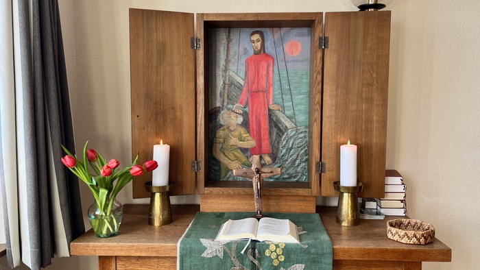 Holzschrank mit geöffneten Türen, der Kunstwerke zeigt, flankiert von Kerzen und Blumen, mit einer Tischdecke und Büchern darunter.