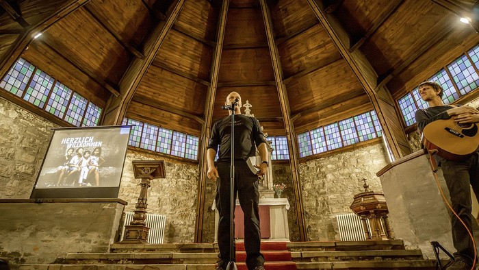 Ein Mann steht auf einer Bühne in einer Kirche und hält ein Mikrofon, während ein anderer Mann hinter ihm eine Gitarre spielt