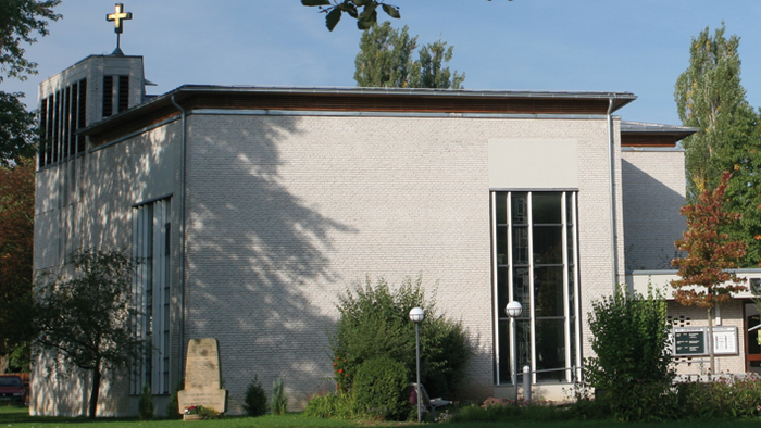 Kleine Kirche mit Kreuz auf dem Dach, umgeben von Bäumen und Gras