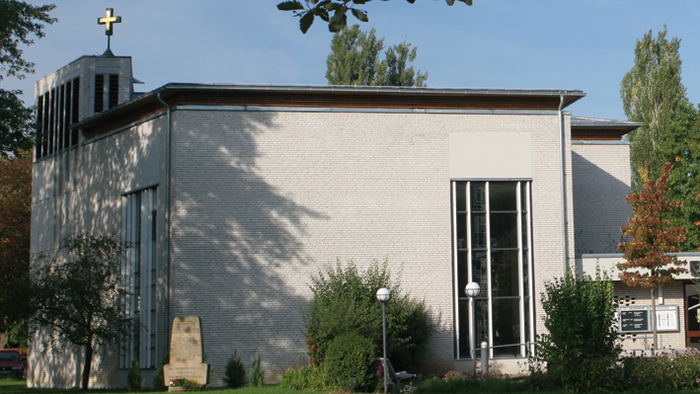 Kleine Kirche mit Kreuz auf dem Dach, umgeben von Bäumen und Gras