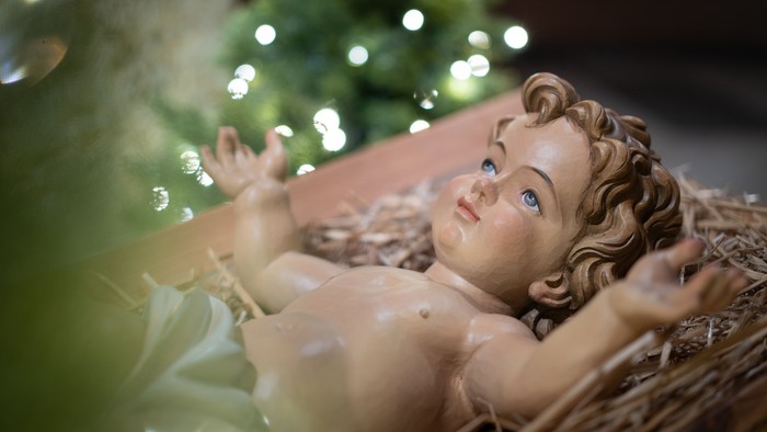 Ein Babyfigur liegt in einem Korb, im Hintergrund ein beleuchteter Weihnachtsbaum.