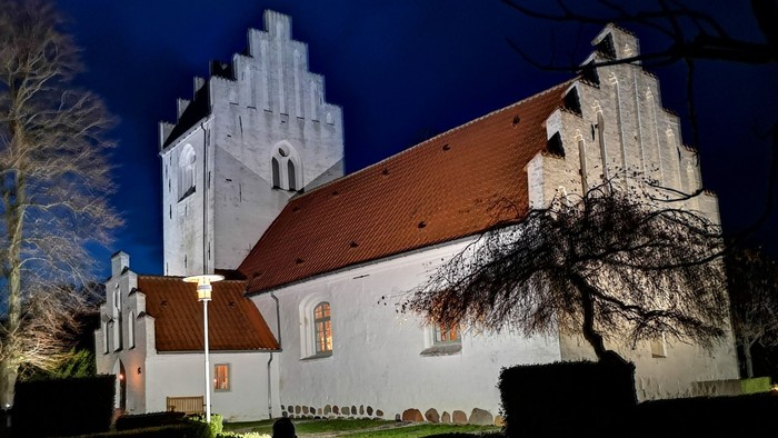 En belyset kirke med tårn og rødt tag omgivet af træer og mørke nat.