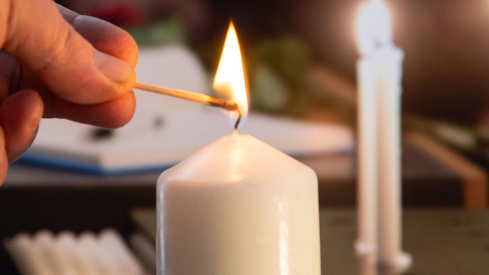 Eine Hand zündet mit einem Streichholz eine weiße Kerze an, während im Hintergrund eine weitere brennende Kerze steht.