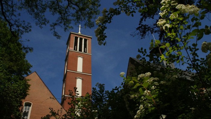 Ein roter Kirchturm ragt zwischen Bäumen und Büschen in den blauen Himmel