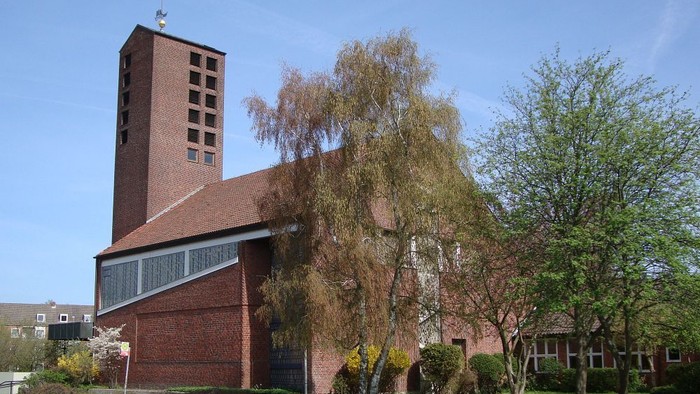 Ein modernes Ziegelgebäude mit hohem Turm und spitzem Dach, umgeben von Bäumen und Büschen.