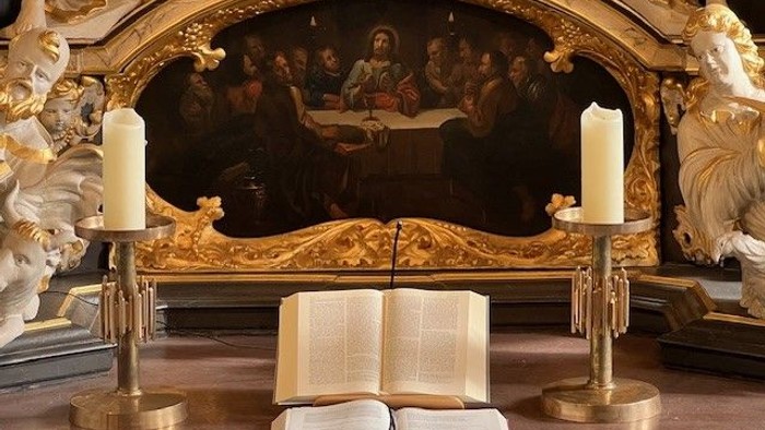 Goldgerahmtes Gemälde mit biblischer Szene, flankiert von zwei Kerzen und einem aufgeschlagenen Buch auf einem Altar.