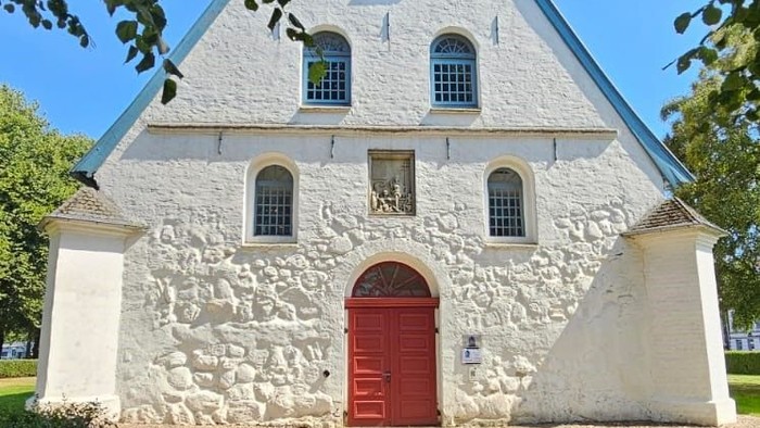 Weißes steinernes Gebäude mit rotem Tor und spitzem Dach unter grünen Bäumen