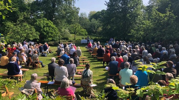 Große Menschenmenge sitzt im Freien auf Bänken und hört einem Redner zu, umgeben von Bäumen.