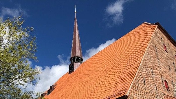 Ein rotes Kirchendach mit spitzem Turm vor blauem Himmel