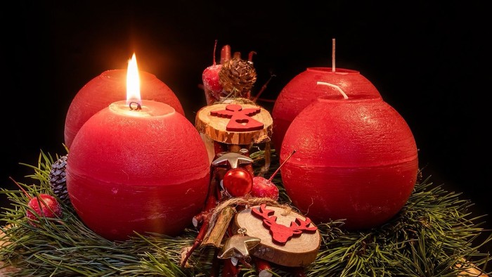 Drei rote Kerzen auf grünem Hintergrund, eine angezündet, umgeben von Weihnachtsdekorationen