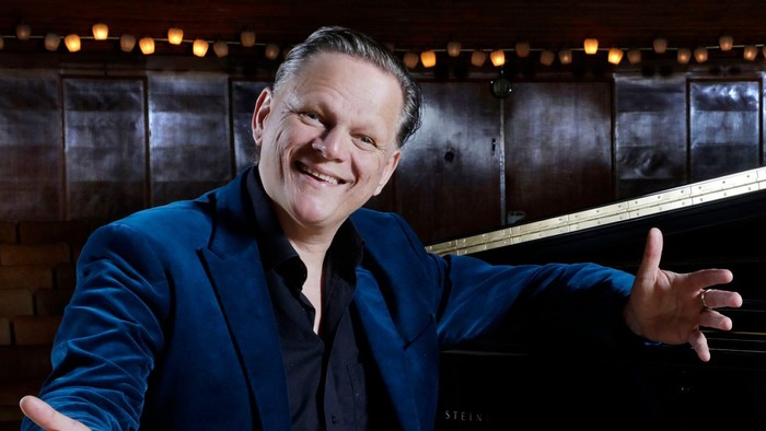 Mand i blå jakke står ved piano og smiler til kamera.