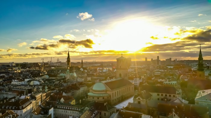 Byen ved solopgang med skyer og tårne.