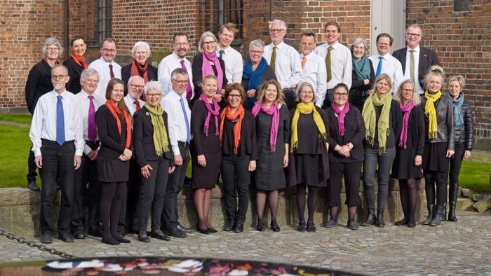 En gruppe af mænd og kvinder står sammen i en række foran en mur af røde mursten. De er alle tildelt med skarves og formelle klæder.