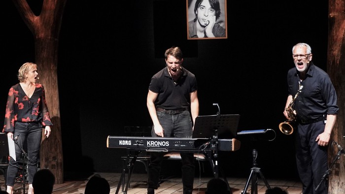 Eine Band spielt auf einer Bühne, ein Mann spielt Saxophon, ein anderer Keyboard, eine Frau singt