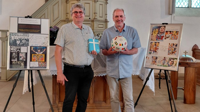 Zwei Männer stehen stolz in einer Kirche mit historischen Schautafeln und halten einen Fußball und Geschenkboxen.