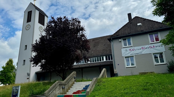 Kirchengebäude mit Uhrturm und Treppe zum Eingang, unter bewölktem Himmel.