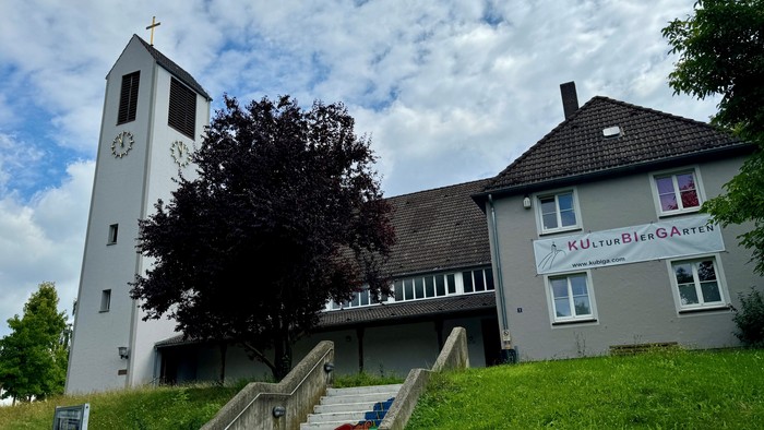 Kirchengebäude mit Uhrturm und Treppe zum Eingang, unter bewölktem Himmel.