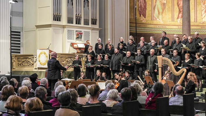 Ein Chor singt in einer Kirche, dirigiert von einem Mann, während ein Orchester spielt und Zuhörer zuhören.
