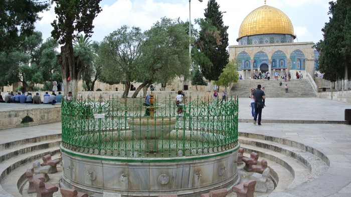 Im Vordergrund ein Brunnen, im Hintergrund eine Kuppelmoschee mit Menschen.