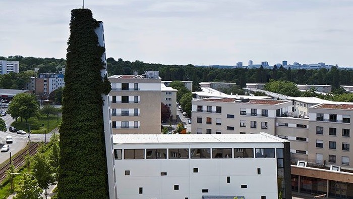 Städtische Szene mit einem hohen Schornstein neben modernen Gebäuden und Grünflächen unter einem bewölkten Himmel.