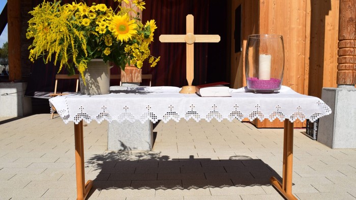 Auf dem Bild sieht man einen Altar im Kurpark vor dem Gradierwerk in Bad Sassendorf. Es steht ein Holzkreuz, eine Kerze in einem Glas und ein Sonnenblumenstrauß auf einem Holztisch, der mit einer weißen Tischdecke gedeckt ist.