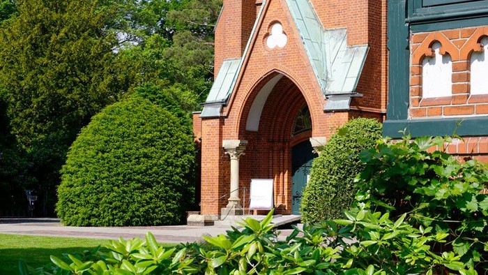 Das Bild zeigt eine rote Ziegelkirche mit grünen Büschen und Bäumen.