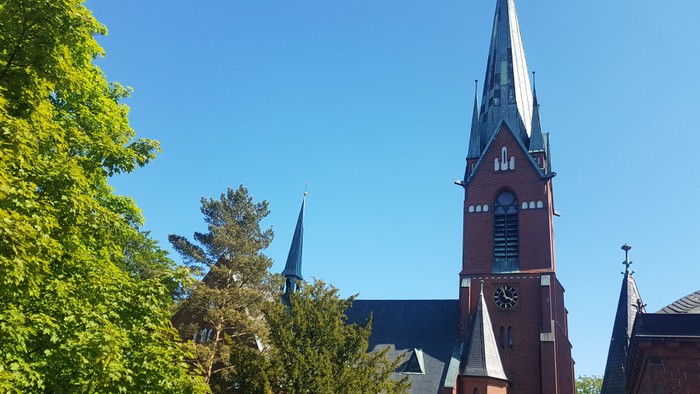 Ein rotes Kirchgebäude mit hohem Turm und Uhr, umgeben von grünen Bäumen unter blauem Himmel