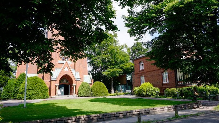 Das Bild zeigt eine rote Backsteinkirche mit hohem Torbogen, umgeben von üppigem Grün und Bäumen.
