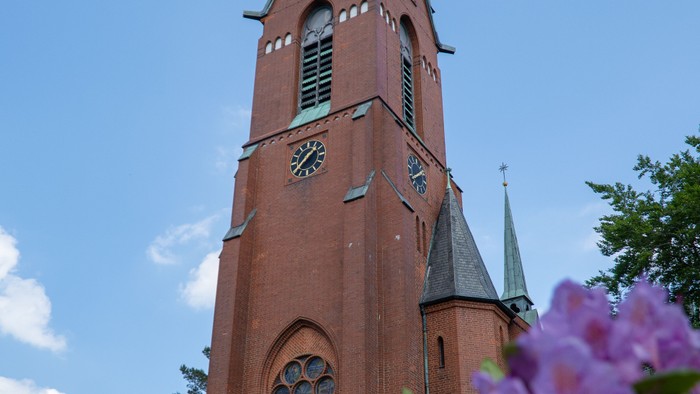 Ein roter Kirchturm mit Uhr und spitzen Dachaufsätzen vor blauem Himmel und lila Blumen im Vordergrund
