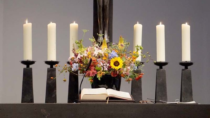 Zwei Reihen von Kerzen flankieren eine Vase mit bunten Blumen auf einem Altar.