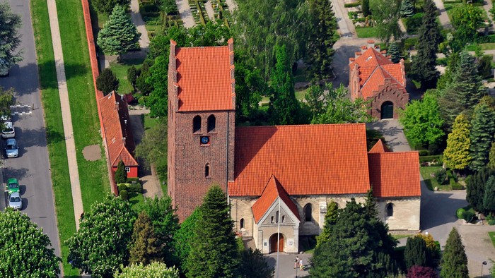 En kirke med røde tag og omgivelser med træer og en parkering.