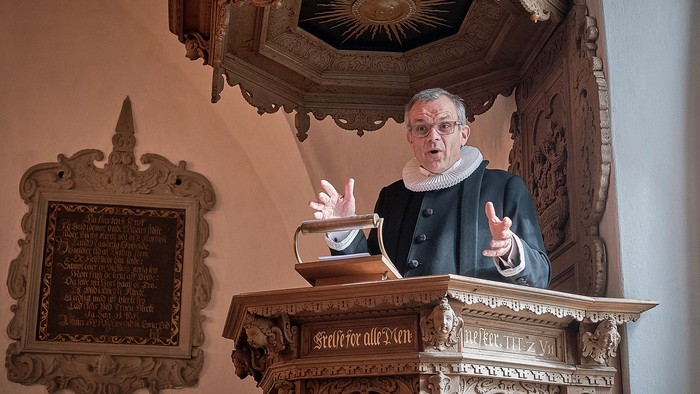 En præst i træstue med et skriftstykke på væggen bagved