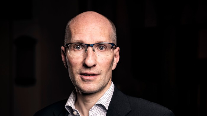 En bald, brillet mand i mørkt jakkesæt og hvid og blå fløjtebluse.