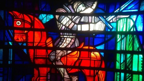 Buntglas-Kunstwerk mit einer roten Figur mit ausgebreiteten Armen, umgeben von blauen und grünen Feldern.