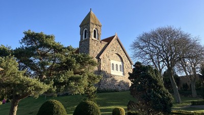 Hyggelig stenkirke med klokketårn omgivet af veltrimmet grønne områder under en klar blå himmel.