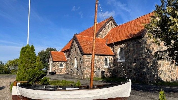 En lille hvid båd med mast foran en stenhus med røde tag.