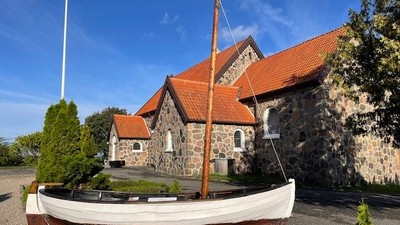 En lille hvid båd med mast foran en stenhus med røde tag.