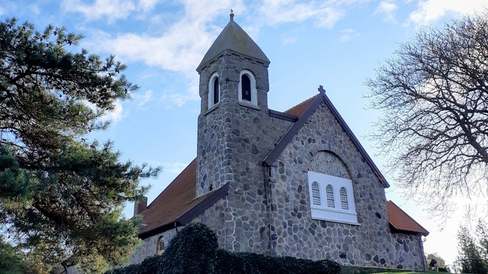 En lille stenkirke med tårn og bjergtagende tag på en bakke med græs og træer.