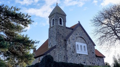 En lille stenkirke med tårn og bjergtagende tag på en bakke med græs og træer.