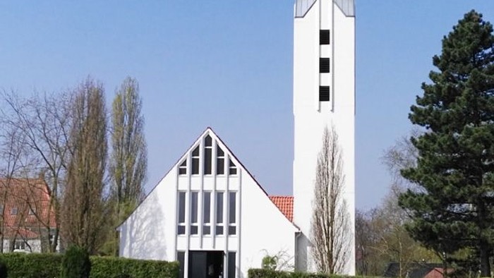 Weiße Kirche mit hohem Turm und spitzem Dach, umgeben von Bäumen und Büschen