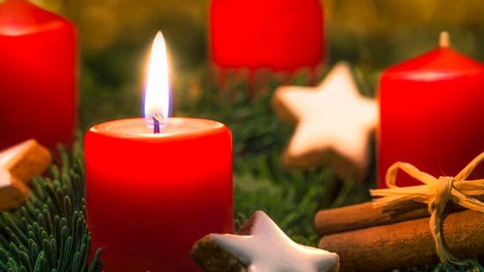 Vier rote Kerzen, drei unangezündet, eine angezündet, umgeben von Tannenzweigen, Zimtstangen und sternförmigen Dekorationen.