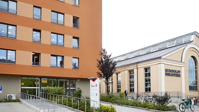 Modernes Gebäude mit orangefarbener Fassade und Glasfront, daneben ein beigefarbenes Gebäude mit Säuleneingang und Fahrrädern davor