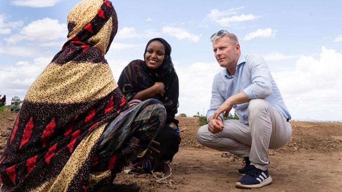 To tre personer sidder på jorden i en åben mark. En mand i blå klæder og to kvinder i traditionelle tøjer.