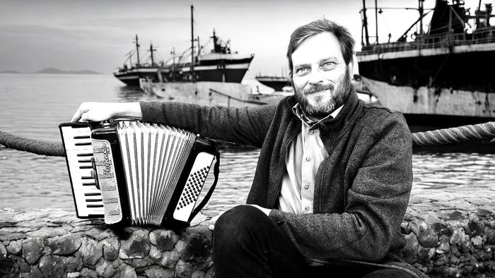 Ein Mann sitzt auf Felsen und spielt Akkordeon, hinter ihm ein Schiff im Hafen.